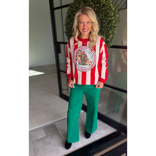 A woman stands in front of a glass door wearing a festive holiday sweater with red and white vertical stripes, a large circular Christmas tree graphic on the front, and bright green wide-leg pants.