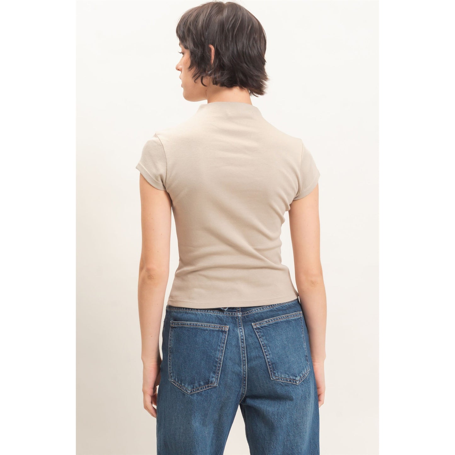 A woman shown from the back wearing a beige short-sleeve mock neck top and blue jeans against a plain off-white background.