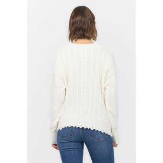 A woman viewed from behind wearing a cream cable-knit sweater and blue jeans against a plain light gray studio background.