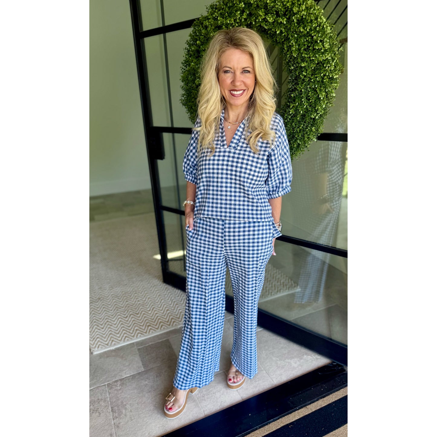 A woman wearing a blue and white gingham short-sleeve jumpsuit with a button-up front and wide-leg pants, standing in front of a glass door with a green wreath.