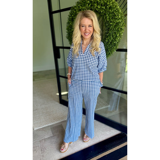 A woman wearing a blue and white gingham short-sleeve jumpsuit with a button-up front and wide-leg pants, standing in front of a glass door with a green wreath.