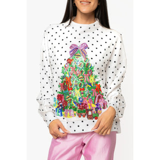 A woman wearing a white crewneck sweatshirt with black polka dots and a colorful Christmas tree graphic on the front, paired with pink pants, against a light gray studio background.