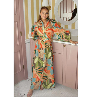 A woman models a long-sleeved maxi shirt dress with a vibrant, multicolor tropical floral print. The dress features a button-up front, a matching fabric tie belt, and a high-low hemline.