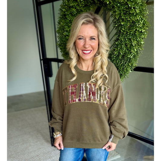 A smiling blonde woman wearing an olive green crewneck sweatshirt with 'TEAMS' in a plaid pattern across the chest, paired with blue jeans, standing in front of a glass door with a green wreath.