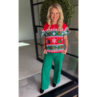 A woman stands in front of a glass door wearing a festive holiday sweater with a red, green, and white Christmas pattern, paired with bright green wide-leg pants and black shoes.