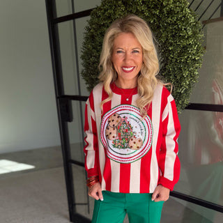 A smiling blonde woman stands indoors wearing a festive red and white vertically striped long-sleeve sweater with a large holiday cookie-and-tree graphic on the front, paired with bright green pants.
