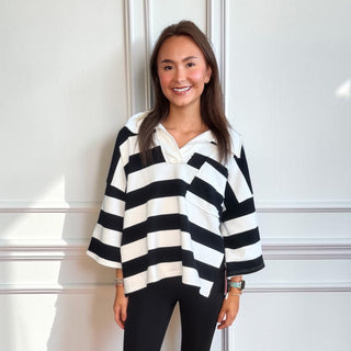 A woman wearing a black and white horizontally striped long-sleeve collared top with black leggings, standing against a white paneled wall.