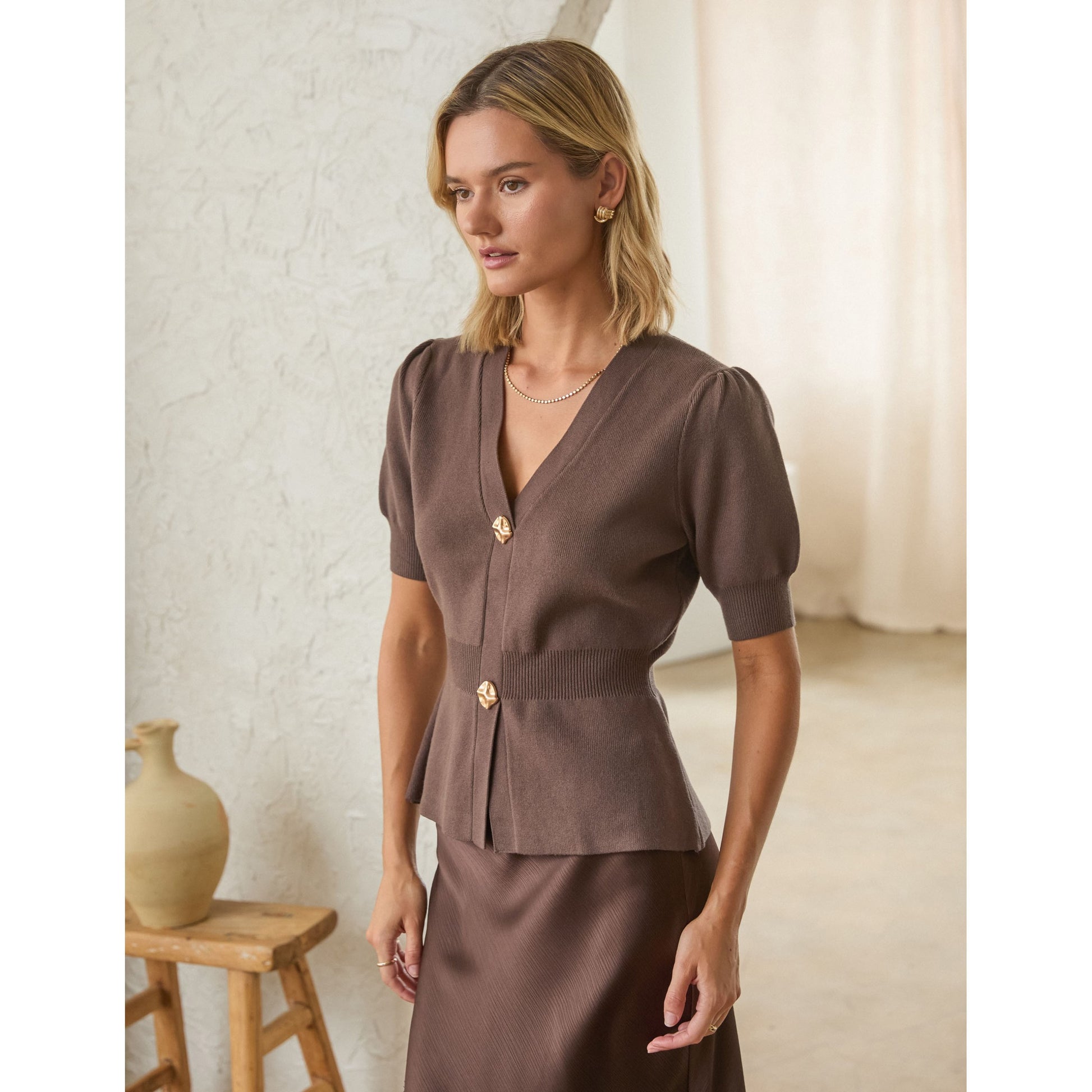 A woman models a brown short-sleeve peplum top with gold buttons, paired with a matching brown skirt, in a softly lit indoor setting.