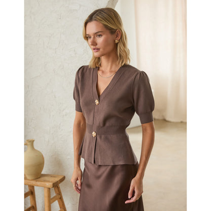 A woman models a brown short-sleeve peplum top with gold buttons, paired with a matching brown skirt, in a softly lit indoor setting.