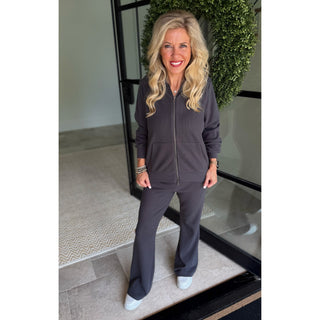 A woman stands indoors wearing a matching dark gray zip-up hoodie and flared pants set, with white sneakers. The photo is taken in front of a glass door with a green wreath behind her.