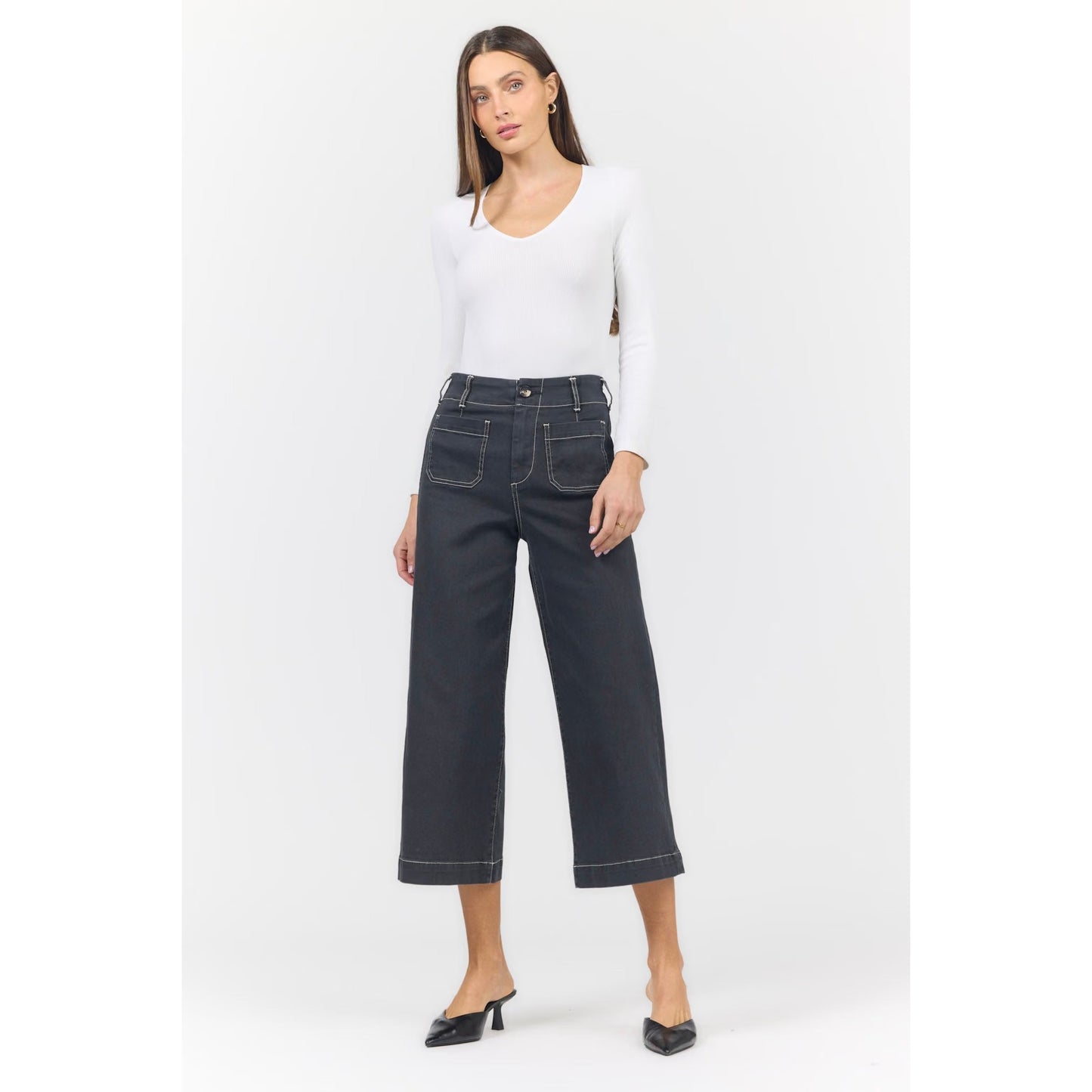 A woman modeling a pair of dark charcoal cropped wide-leg jeans with white contrast stitching, paired with a white long-sleeve top and black pointed-toe mule heels, against a light gray studio background.