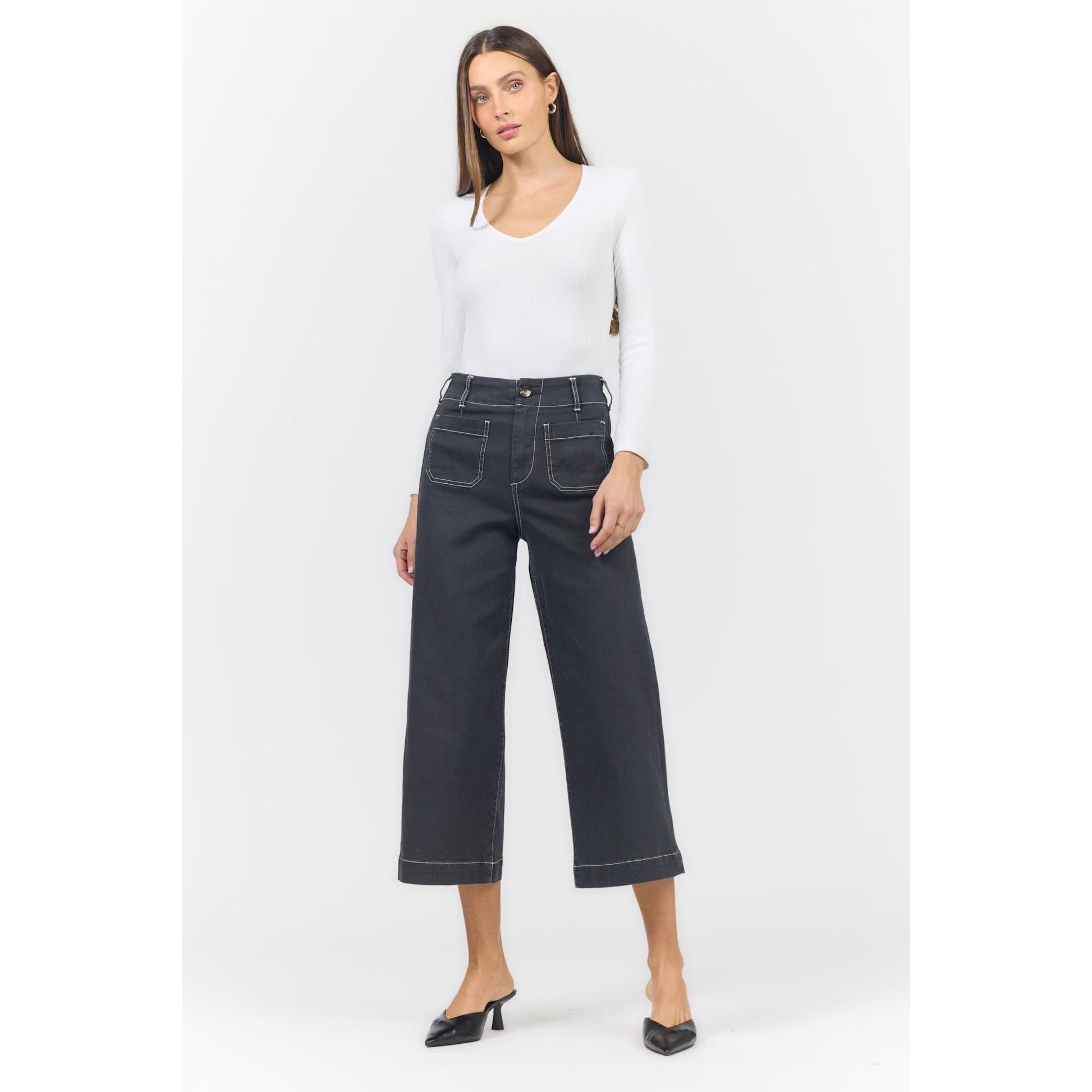 A woman modeling a pair of dark charcoal cropped wide-leg jeans with white contrast stitching, paired with a white long-sleeve top and black pointed-toe mule heels, against a light gray studio background.