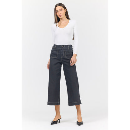 A woman modeling a pair of dark charcoal cropped wide-leg jeans with white contrast stitching, paired with a white long-sleeve top and black pointed-toe mule heels, against a light gray studio background.