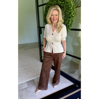 A woman modeling a cream short-sleeve peplum top and brown wide-leg pants, standing in a modern entryway with a green hedge and glass door visible.