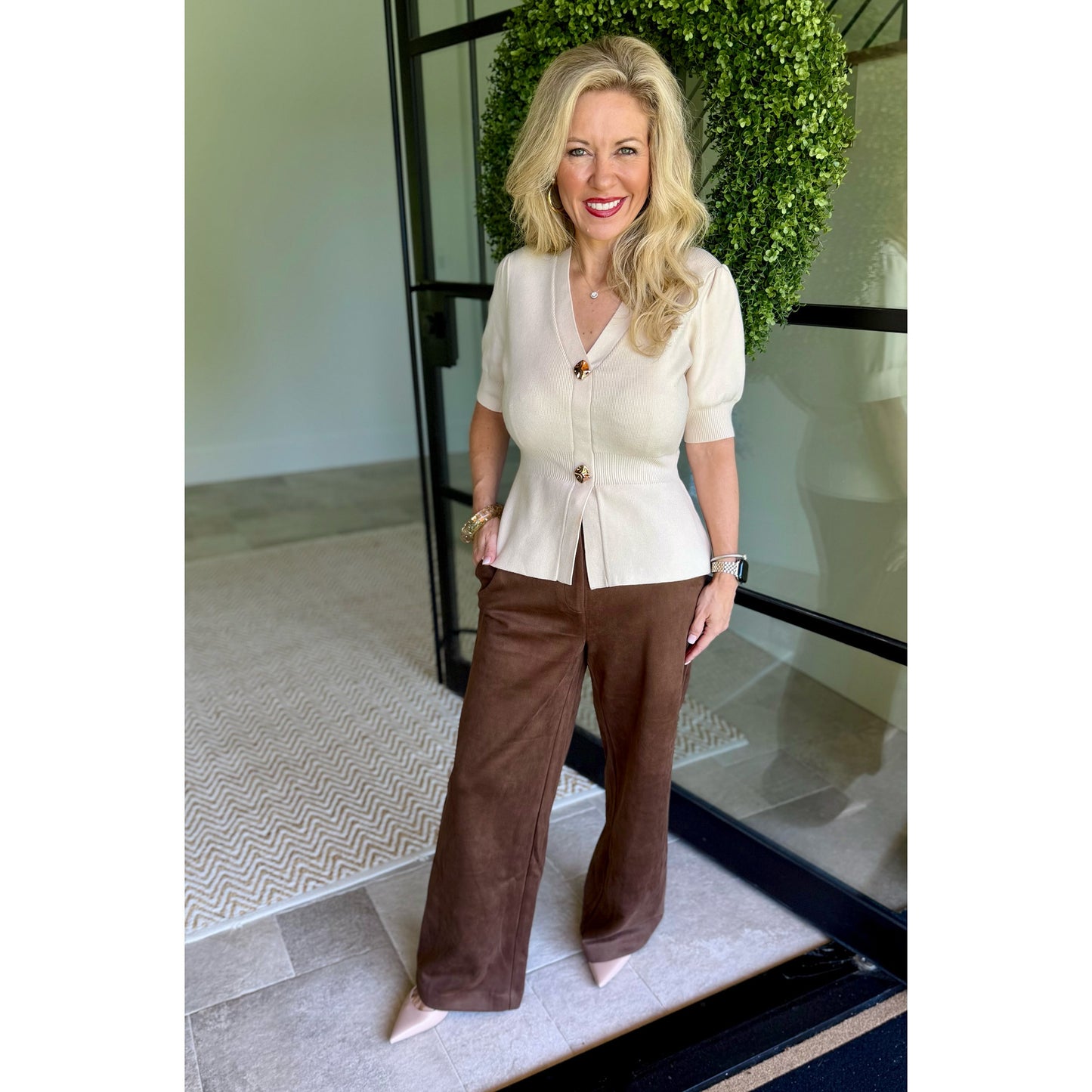 A woman modeling a cream short-sleeve peplum-style top with brown buttons and matching brown wide-leg pants, standing indoors near a glass door and greenery.