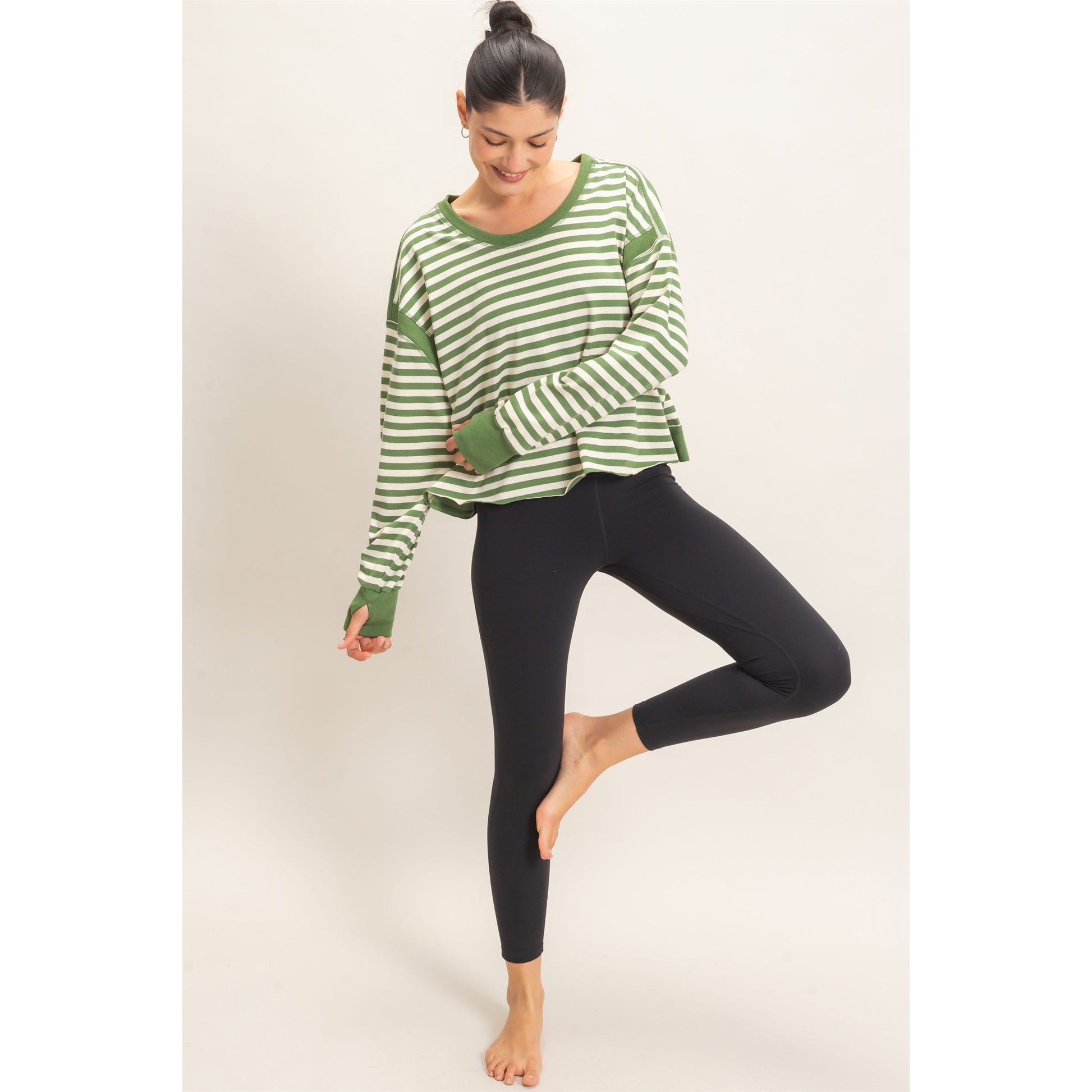 A woman modeling a green and white striped long-sleeve top with black leggings, standing in a yoga tree pose against a plain light beige studio background.