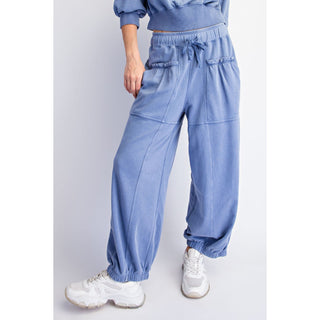 A person wearing loose, wide-leg blue jogger-style pants with elastic cuffs, a drawstring waist, and front patch pockets, paired with white sneakers against a light gray studio background.