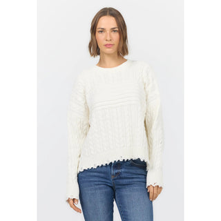 A woman modeling a cream-colored cable-knit sweater with a scalloped hem and distressed details on the sleeves, paired with blue jeans, against a light gray studio background.