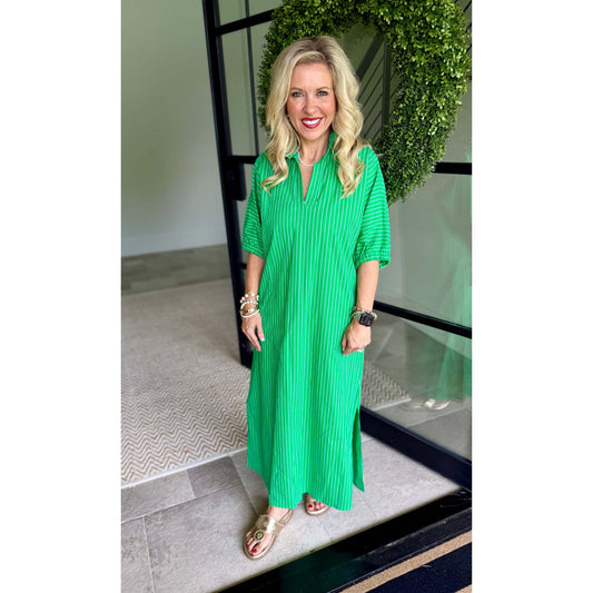 A woman wearing a bright green pleated midi dress with short sleeves and a split neckline, standing in front of a glass door with a green wreath in the background.