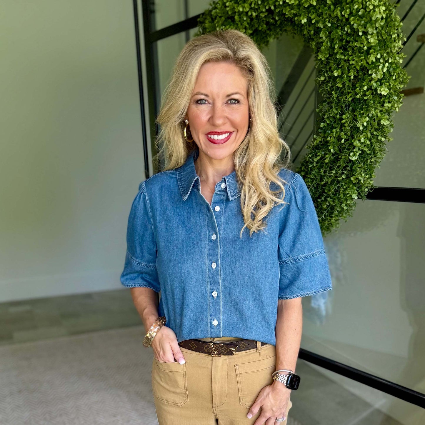 A smiling blonde woman wearing a short-sleeve blue denim button-up shirt and tan pants, standing indoors in front of a glass wall with a green leafy wreath.