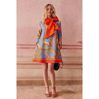 A woman models a light blue silk dress with a vibrant orange and yellow abstract pattern and a large matching orange bow at the collar, paired with orange heels and a small dark blue handbag, set against a pinkish-beige stone wall.