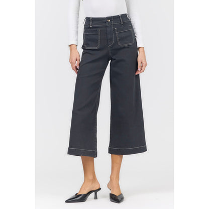 A model wearing a pair of dark charcoal gray cropped wide-leg denim jeans with white contrast stitching, paired with a white long-sleeve top and black pointed mule heels, against a light gray studio background.