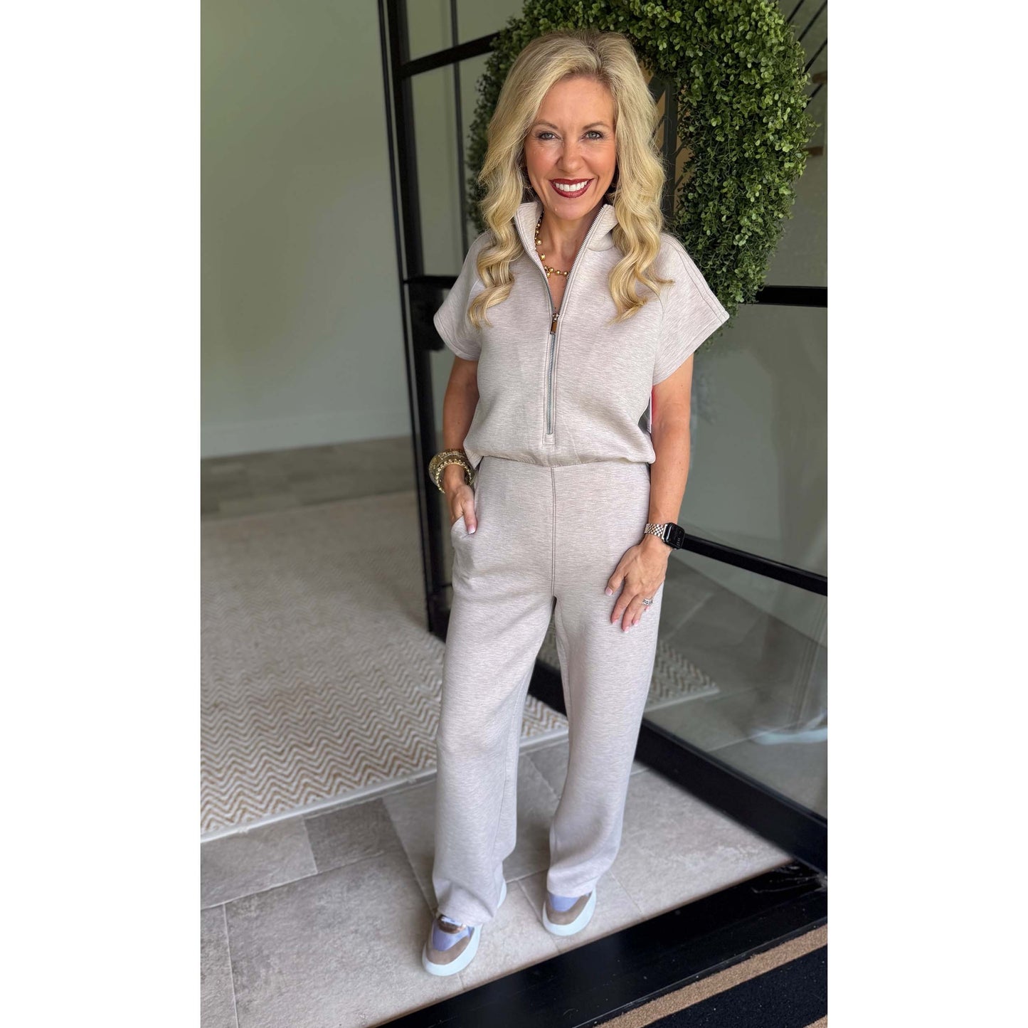 A smiling blonde woman stands indoors wearing a light beige short-sleeve zip-front jumpsuit with wide-leg pants and white sneakers. The photo is taken in a modern interior with a glass door, black frame, and greenery in the background.