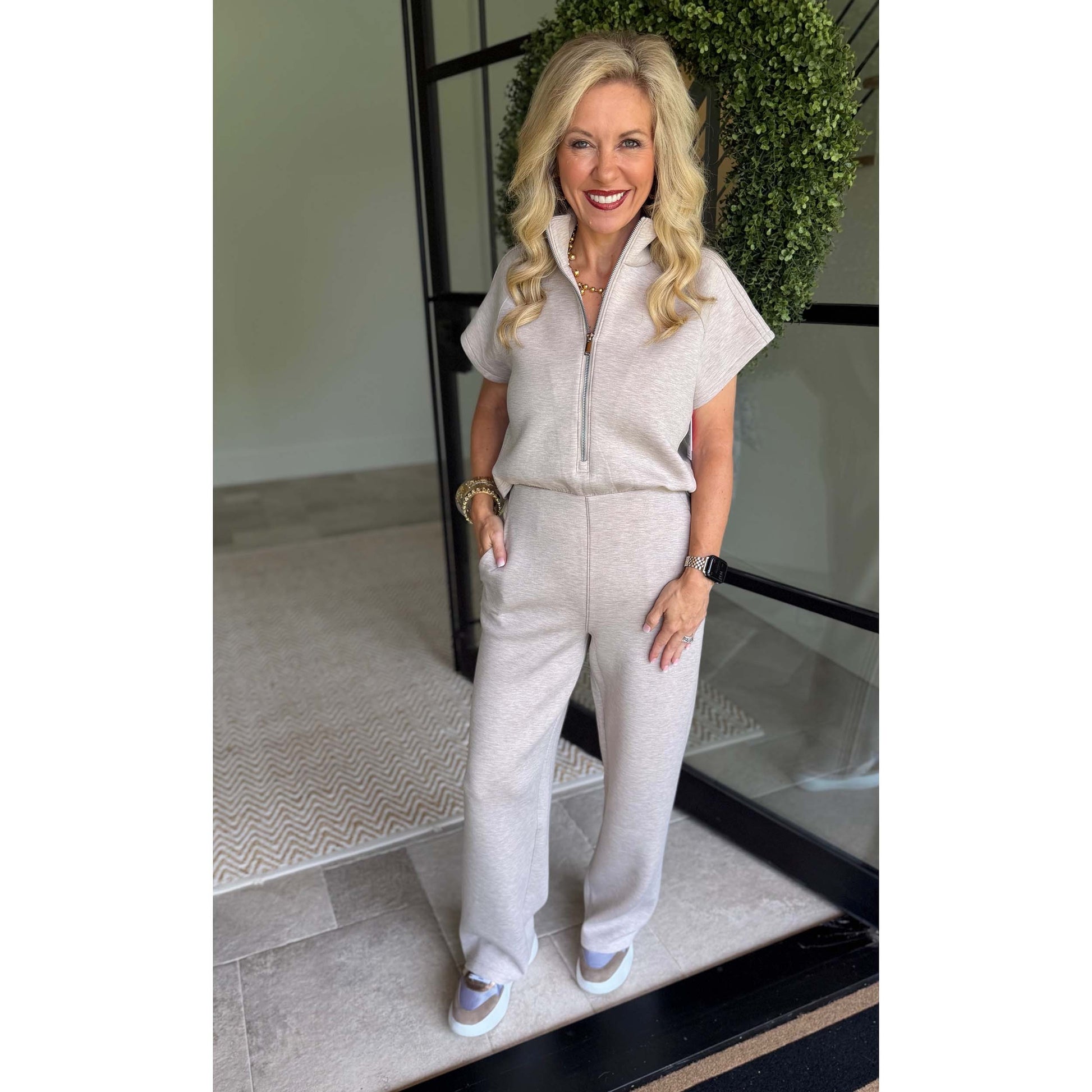 A smiling blonde woman stands indoors wearing a light beige short-sleeve zip-front jumpsuit with wide-leg pants and white sneakers. The photo is taken in a modern interior with a glass door, black frame, and greenery in the background.
