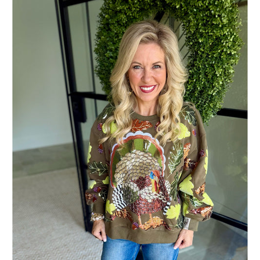 A smiling blonde woman wearing a brown crewneck sweatshirt with a large sequined turkey design and autumn leaf patterns, paired with blue jeans, standing in front of a green leafy wall.