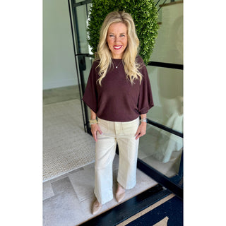 A woman stands indoors wearing a loose short-sleeve top in dark plum-brown and cream wide-leg pants, with beige heels. She is posed in front of a glass door with a green hedge behind her.