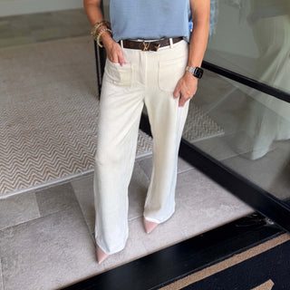 A person wearing off-white wide-leg trousers with a brown leather belt, paired with a light blue top and pink pointed-toe heels, standing on a tiled floor.