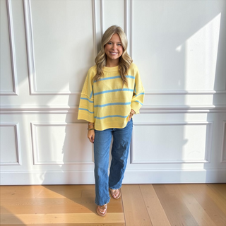 A woman stands in a bright room with white paneled walls and light wood floors, wearing a yellow knit sweater with light blue horizontal stripes and blue jeans.
