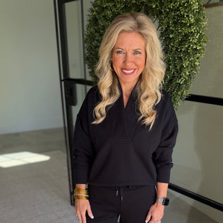 A smiling blonde woman wearing a black long-sleeve mock neck top and matching black pants, standing indoors in front of a glass door and a green leafy wall.
