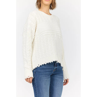 A woman wearing a cream-colored cable-knit sweater with a frayed hem and cuffs, paired with blue jeans, shown from the neck down against a plain white background.