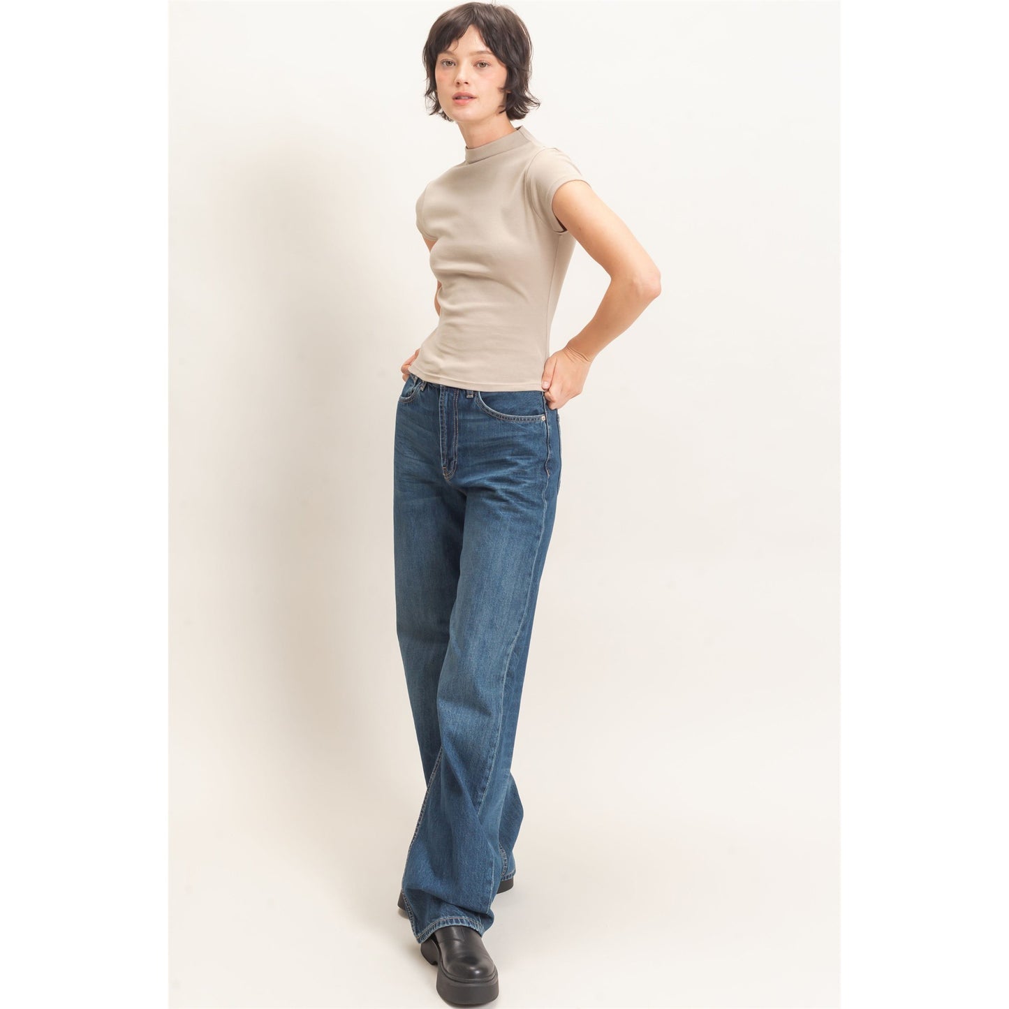A model wearing a beige short-sleeve mock neck top and blue wide-leg jeans, standing against a plain off-white studio background.
