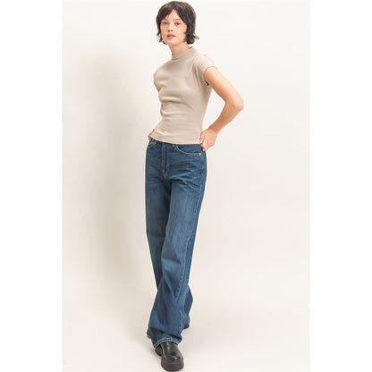 A model wearing a beige short-sleeve mock neck top and blue wide-leg jeans, standing against a plain off-white studio background.